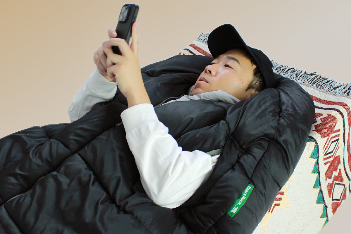 寝袋から手を出してスマホを見る男性